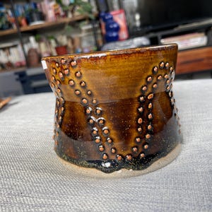 Puede incluir: Una taza de cerámica marrón con un esmalte moteado y un patrón de pequeños puntos marrones. La taza tiene una base ancha y un borde ligeramente acampanado.