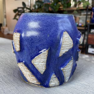May include: A handmade ceramic vase with a deep blue glaze and textured, cream-colored geometric cutouts. The vase has a cylindrical shape and is suitable for displaying flowers or as a decorative item.