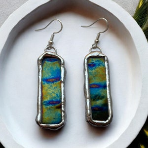 May include: A pair of silver earrings with iridescent blue, green, and yellow glass inserts. The glass is encased in a silver frame with a slightly irregular shape.
