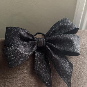 May include: A black glitter bow hair accessory. The bow is made of a shiny, textured fabric and is tied with a black elastic band. The bow is a classic shape with multiple loops and tails, perfect for adding a touch of sparkle to any hairstyle.