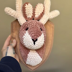 May include: A plush, crocheted deer head mounted on a wooden plaque. The deer features a brown and white face, with tan antlers and ears. The deer head is a decorative item, suitable for a nursery or rustic decor.