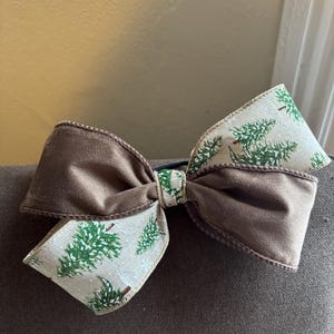 Könnte beinhalten: Eine große braune Haarschleife mit einem Weihnachtsbaum-Muster. Die Schleife hat eine durchgehend braune Mitte und Ränder, mit einem cremefarbenen Band mit grünen Weihnachtsbäumen bedruckt. Die Schleife ist mit einem schwarzen Gummiband gebunden.