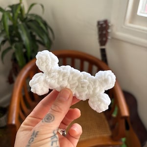 May include: A handmade, white crochet bone-shaped toy. The plush toy is held in a hand, with a wooden chair and a guitar in the background. The toy is made of soft, fluffy yarn.