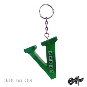Puede incluir: Llavero con la letra V verde con la palabra "VEGAN" escrita en letras blancas en la parte delantera. El llavero está unido a una cadena plateada y un anillo plateado.