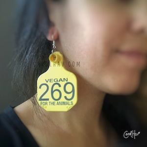 May include: Yellow vegan earrings with the text "VEGAN 269 FOR THE ANIMALS". The earrings are shaped like a tag and have a silver hook. The earrings are a statement piece.