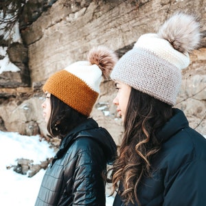 May include: Two people wearing winter jackets and knit hats with pom poms. The hats are white, brown, and beige. The jackets are black.