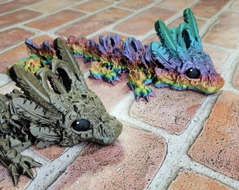 May include: Two articulated dragon figures. One is a muted gray-green, the other is a vibrant rainbow of colors. Both have detailed feather-like features and large, dark eyes. The dragons are posed on a brick surface, showcasing their intricate design.