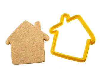 Puede incluir: Un cortador de galletas de plástico amarillo con forma de casa, junto a una galleta sencilla de color marrón claro cortada en la misma forma.