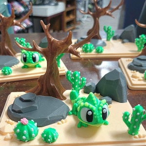 May include: A 3D printed desert diorama featuring a bright green chameleon with large eyes, a brown leafless tree, grey rocks, and several green cacti with white spots on a tan rectangular base. One small cactus has a pink flower on top.