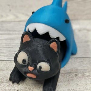 May include: A 3D printed figurine featuring a bright blue shark with a white-toothed open mouth positioned behind a black cat. The cat has large white eyes, a small pink nose, and pink inner ears, appearing to be caught inside the shark's mouth.