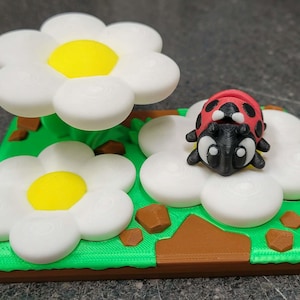 May include: A 3D printed decorative figurine featuring a red and black ladybug resting on a white flower with a yellow center. The scene includes a second white flower, green grass, and brown rock accents on a rectangular base with a brown border.