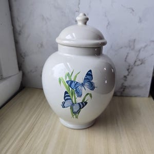 May include: A cream-colored ceramic jar with a lid, featuring a butterfly design. The jar has a rounded shape and a small knob on top. The butterflies are blue and white, with green leaves.