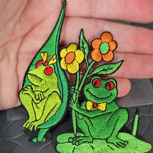 May include: Embroidered patches featuring cartoon frogs. One frog is inside a green leaf, the other holds flowers. Both frogs are green with red eyes and yellow accents. The patches are designed with detailed stitching.