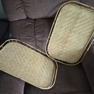May include: Two rectangular serving trays constructed from light tan woven bamboo with a natural finish. Each tray features a sturdy bamboo frame with a raised border and a flat, tightly woven base, showcasing a consistent, repeating basket-weave pattern.