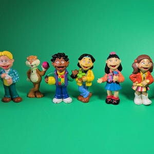 May include: A set of eight colorful plastic character figurines featuring a brown dog, a boy in a blue shirt, a tan animal, a boy in a colorful vest, a person in a yellow jacket, a girl in a blue skirt, a girl in an orange jacket, and a person in a purple hat.