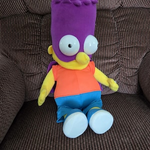 May include: A plush Bart Simpson doll in a superhero costume. The doll features a purple mask and head covering, an orange shirt, blue pants, and large white eyes. The doll is sitting on a brown patterned surface.