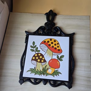 May include: A black cast iron trivet with a ceramic tile featuring a colorful mushroom design. The mushrooms have red, orange, and brown caps with yellow spots, set against a white background. The trivet has a decorative handle.