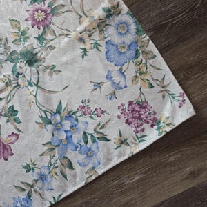 May include: White floral tablecloth featuring a repeating pattern of blue, purple, and yellow flowers with green leaves. The tablecloth is made of a textured fabric and is set against a dark wood surface.