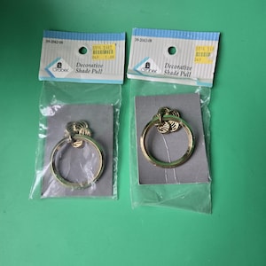 May include: Two decorative shade pulls in clear plastic packaging. Each pull has a gold-colored ring with a decorative top. The packaging includes the text "Decorative Shade Pull" and the brand name "Grober".