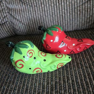 May include: Two ceramic chili pepper figurines. One is red with black triangles and white dots, the other is green with red swirls and black dots. Both have green stems.