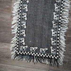 May include: A black and white woven textile runner featuring a central pattern of small dots, bordered by a repeating cow motif and a checkered edge. The piece is finished with long, white fringe along the sides and bottom edges.