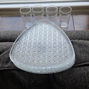 May include: A clear glass triangular serving tray with a textured, starburst pattern, stacked with four matching clear drinking glasses. The glasses are cylindrical with a slightly flared top. The items are on a dark brown surface.