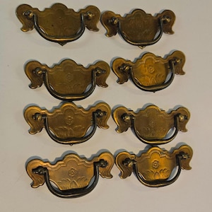May include: A set of eight brass-toned metal drawer pulls, each featuring a decorative backplate with an embossed floral motif and a curved, swinging bail handle. The hardware displays a weathered, aged finish with intricate detailing.