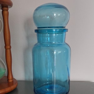 May include: A clear, blue glass apothecary jar with a rounded lid. The jar has a cylindrical body and a wide mouth. The lid is a rounded shape. The jar is sitting on a gray surface.