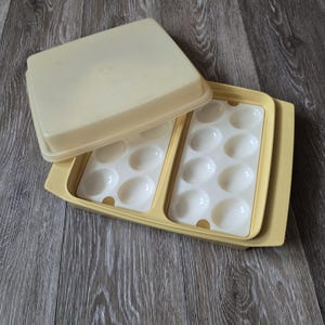 May include: A vintage egg storage container with a cream-colored lid and base. The container has two trays, each with oval-shaped compartments to hold eggs. The lid is slightly ajar, revealing the egg compartments. The container is made of plastic.
