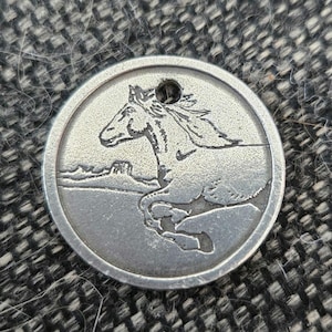 May include: A silver-colored round pendant with a running horse design. The horse is depicted in profile, with a landscape in the background. The pendant has a small hole at the top for a chain or cord.