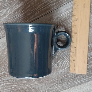 May include: A dark teal ceramic mug with a rounded handle. The mug is approximately 3 inches tall and has a glossy finish. A wooden ruler is placed next to the mug for scale.