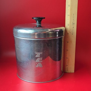 May include: A vintage stainless steel tea canister with a black handle. The canister is cylindrical, with the word "TEA" embossed on the front. The canister is approximately 5 inches tall.