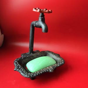 May include: A black cast iron soap dish shaped like a faucet with a red handle. The dish holds a green bar of soap. The soap dish has an ornate, decorative design and sits on a red background.