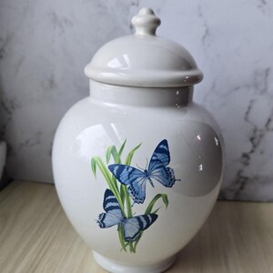 May include: A white ceramic jar with a lid, featuring a blue butterfly design. The jar has a rounded shape and a glossy finish. The butterflies are depicted with detailed wings and are set against green foliage.