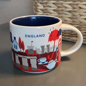 May include: White ceramic mug with a blue interior and a wraparound design featuring iconic England imagery. The design includes a red umbrella, a telephone booth, and the word "ENGLAND".