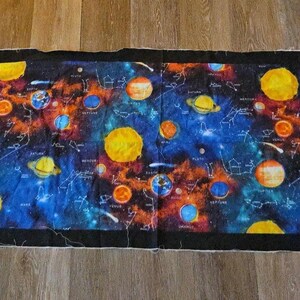 May include: A rectangular fabric panel featuring a vibrant space-themed print with various planets, stars, and constellations set against a deep blue and black nebula background. The design is framed by a solid black border on all four sides.