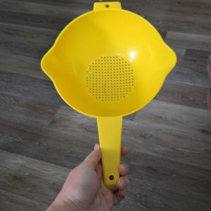 May include: A bright yellow plastic colander with a handle. The colander has a round bowl shape with small holes for draining. The handle has a hole for hanging. The colander is held in a hand.