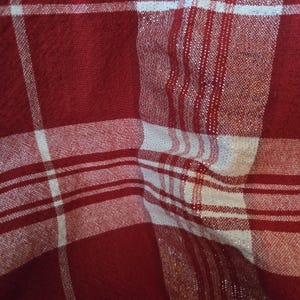 May include: Close-up of a red and white plaid fabric. The fabric has a woven texture with a pattern of intersecting lines, creating a grid-like design. Some sections have a shimmering, metallic thread woven into the fabric.
