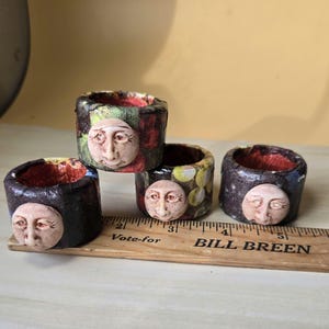 May include: Four handmade napkin rings with a celestial theme. Each ring is cylindrical, with a textured exterior in various colors and patterns. A carved face of the moon adorns each ring. The interior of each ring is red.