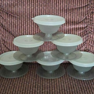 May include: A stack of seven clear plastic dessert cups with white lids. The cups are on clear bases and arranged in a pyramid shape. The background is a patterned brown and pink fabric.