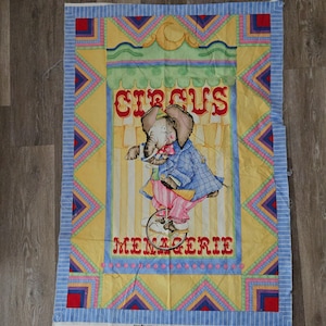 May include: A rectangular fabric panel featuring a circus theme. The central image depicts an elephant riding a unicycle, dressed in a blue coat and pink pants. The word "CIRCUS" is at the top, and "MENAGERIE" at the bottom.