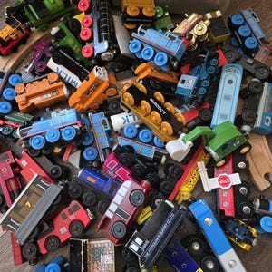 May include: A collection of colorful wooden toy trains, including Thomas the Tank Engine and other characters. The trains are various colors, including blue, red, green, and orange, with black wheels. Some have text like "MURDOCH" and "MAVIS".