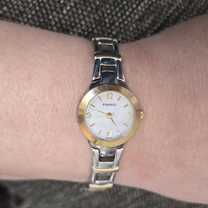 May include: A Fossil brand wristwatch with a gold-tone bezel and silver-tone band. The watch face is white with gold hands and hour markers. The watch displays the date and the time.