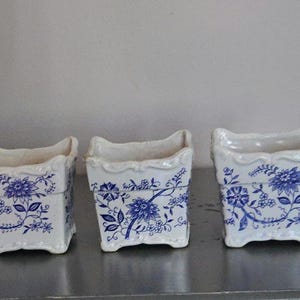 May include: Three square ceramic planters featuring a white base with a blue floral pattern. Each planter has a scalloped top edge and four small feet at the base. The design includes detailed blue botanical illustrations of flowers and vines.