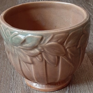 May include: A tan-colored ceramic planter with a decorative leaf pattern around the top. The planter has a rounded shape with vertical ridges and a small, circular base. The rim of the planter has a light green accent.