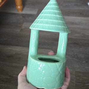 May include: A light green ceramic wishing well figurine with a conical roof and a textured stone-like base. The structure features an open-air design with two vertical supports connecting the base to the roof, creating a hollow center opening.