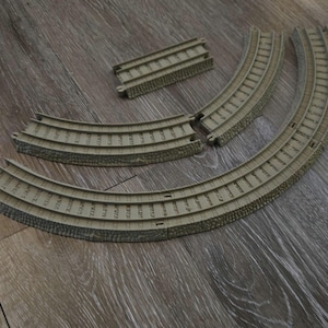 May include: A collection of tan plastic toy train tracks, including curved and straight sections. The tracks are designed to connect and form a railway system. The image shows the tracks on a wooden surface.