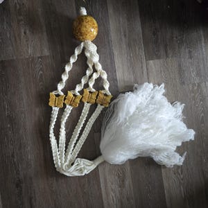 May include: A white macrame plant hanger featuring a large, speckled yellow spherical bead at the top and four smaller, golden-yellow butterfly-shaped beads integrated into the twisted cord design, finished with a long, thick white yarn tassel.