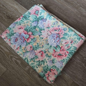 May include: Folded floral fabric featuring a pattern of pink, blue, and yellow flowers with green leaves. The fabric is folded in a square shape, showcasing the detailed floral design. The background is a light cream color.