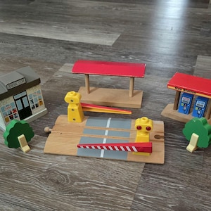May include: Wooden toy train set with a general store, gas station, and railroad crossing. The set includes a red-roofed gas station with blue pumps, a general store, and a crossing with yellow signals and a red and white striped gate.